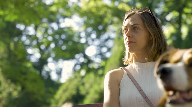 A young woman sits pensively on a wooden bench in a leafy park, framed by the blurred snout of her border collie and a golden retriever beside her. The slow motion tilt shot emphasizes thoughtful pet 