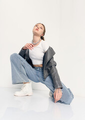 A stylish woman in trendy jeans and a jacket poses sitting on the floor