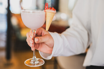 Close-up of a woman's hand holding a pink cocktail in a glass, garnished with a tiny ice cream cone...