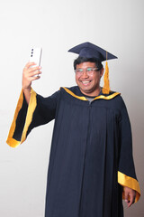 Young man dressed in cap and gown to celebrate his graduation uses his cell phone to take selfies,...