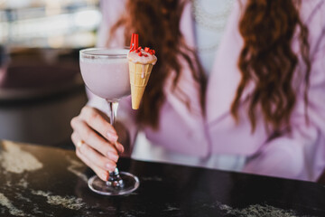 Stylish woman in pink outfit holding a cocktail garnished with a pink cream-topped ice cream cone and red sprinkles, showcasing a creative and elegant drink presentation.