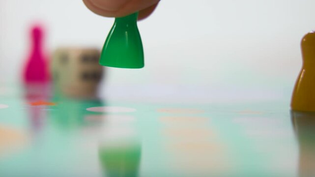 A single green plastic pawn rests on a pastel-colored game board before a hand reaches in to lift and move it, highlighting tactile interaction and detailed macro movement in a smooth steady slow moti