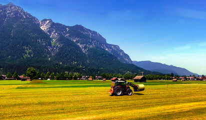 A red tractor plows an agricultural field against the background of forests and mountains on a sunny day.
