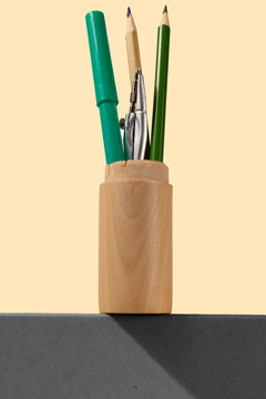 Pencils, a marker, and a compass in a standing wooden holder on a pastel yellow background. The holder stays on top of the grey elevation.