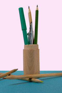 Pencils, a marker, and a compass in a standing wooden holder on a pastel pink background. More pencils lays on the blue desk.