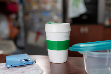 White portable cup with coffee and smartphone. The scene includes everyday item as aplastic container on a wooden surface in kitchen.