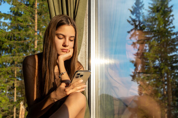Teen girl using smartphone by window reflection. Teenage girl sits by a window with natural light,...