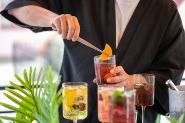 Bartender in black robe uses tweezers to garnish cocktail with orange slice. Colorful drinks and...