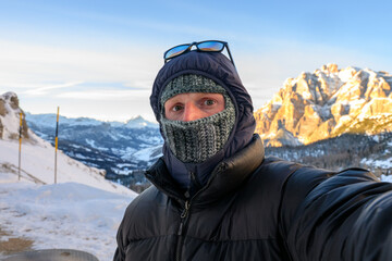 A man wearing a black jacket and a balaclava is taking a selfie in a snowy mountain range
