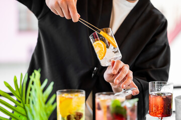 Bartender using tongs to add orange slices into a glass with ice, surrounded by vibrant cocktails....