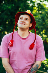 A cheerful man stands in a vibrant outdoor setting wearing a playful hat with a red and yellow...