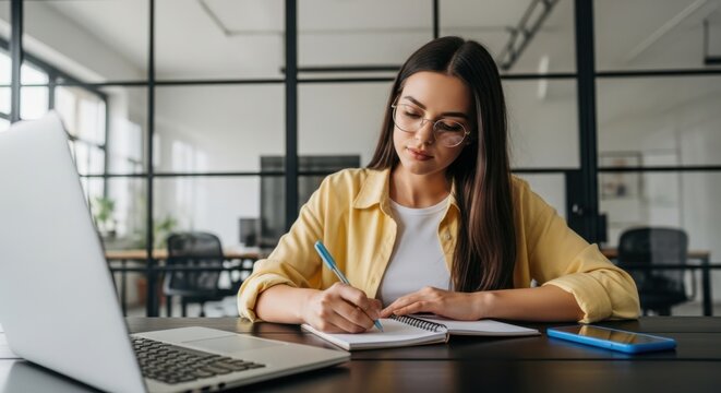 Young woman with glasses is writing in a notebook at her desk - Powered by Adobe