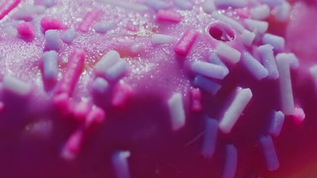 Close-up of a vibrant donut decorated with pink icing and colorful sprinkles glistening under bright lights The rich textures and colors highlight its appetizing appeal