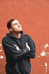 A man in a dark hoodie stands confidently against a weathered orange wall The overcast sky casts a...