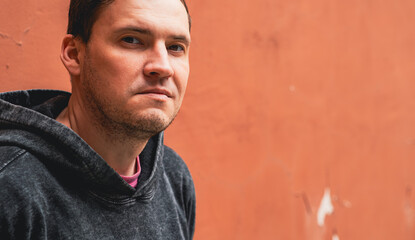A man in a dark hoodie stands confidently against a weathered orange wall The overcast sky casts a...