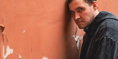 A man stands against a textured wall with peeling paint wearing a dark hoodie His serious...