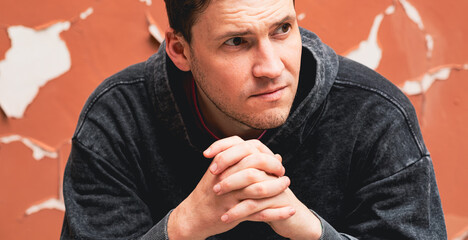 A man wearing a gray hoodie appears lost in thought as he sits against a faded orange wall The wall...
