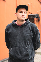 A young man stands confidently in front of a vibrant orange wall wearing a hoodie and cap The urban...