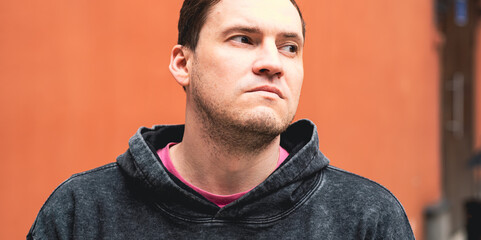 A man with a serious expression stands outdoors wearing a casual hoodie The backdrop features a...