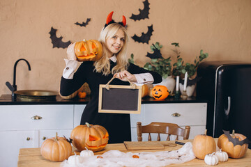 Woman wearing witch costume holding carved pumpkin and blank blackboard in Halloween decorated...