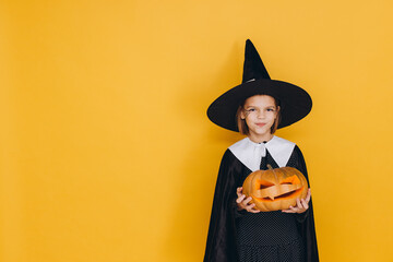 Young girl wearing witch costume holding jack o' lantern posing on yellow background