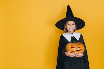 Smiling girl wearing witch costume holding jack o' lantern posing on yellow background for Halloween