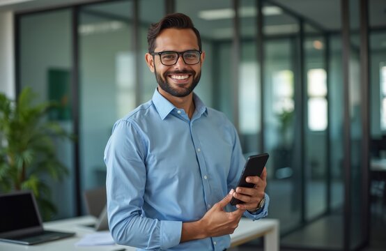 Smiling young Latin business man in office holds phone. Professional executive or employee looks at camera with smartphone. Happy confident businessman at work in modern office. Business concept.