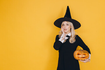 Surprised witch holding jack o' lantern posing on yellow background