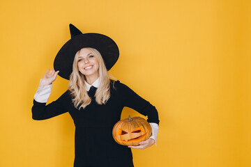 Smiling witch holding jack o' lantern posing on yellow background for halloween party