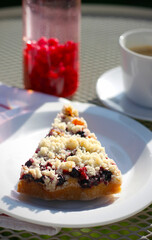 Slice of homemade red and black currant crumb cake on a white plate, with berry lemonade and cup of coffee in background, summer dessert concept, outdoor breakfast or garden cafe moment
