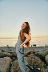 smiling woman in denim outfit enjoying sunset by the sea with natural rocks and calm water background, warm lighting and relaxed summer vibe