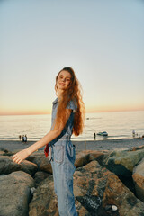 Lifestyle woman in modern boho-western denim street style enjoys sunset by the rocky beach, featuring warm film color tones and relaxed coastal vibe.