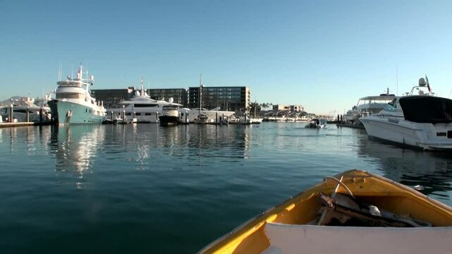 A small yellow boat departs the dock, leaving behind luxurious yachts, modern buildings, and calm waters under a bright, clear sky in Cabo San Lucas, Mexico, during daylight hours.