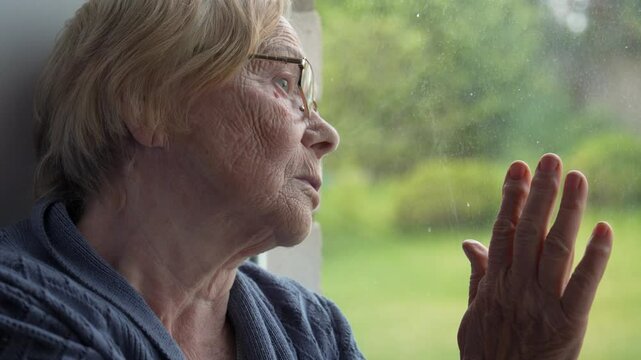Old ill woman wearing glasses. Touching window glass while gazing thoughtfully outside. Elderly female with absent look has dementia or mental illness. Eyes are bulging with fear of being lonely.
