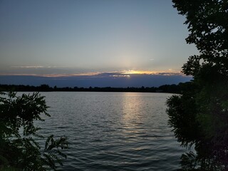 Beautiful sunset over a calm lake with gentle ripples, silhouetted trees framing the scene, and a...