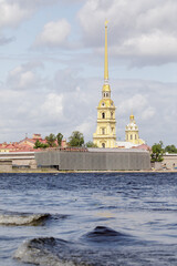 Golden spire of Peter and Paul Cathedral rises above fortress walls, viewed from across the Neva River under cloudy summer skies.