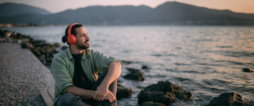 A young man is sitting on the beach with headphones on at sunset by the sea. - Powered by Adobe