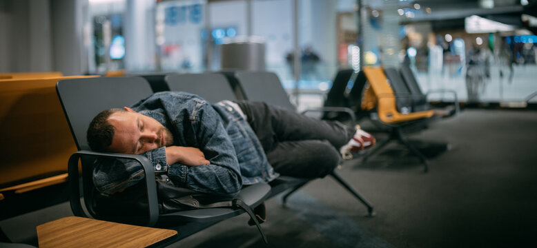 A tired male passenger sleeps on the seats waiting for boarding in the departure area of the airport terminal.