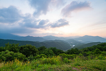 A scenic mountain view in South Korea at sunrise, featuring dramatic clouds and lush summer greenery in the foreground.