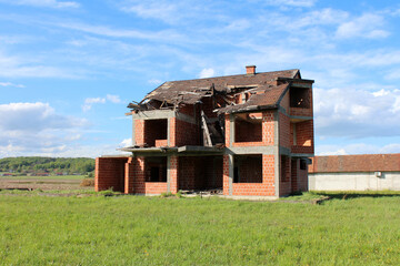 A partially constructed brick house with a collapsed wooden roof stands abandoned in a green field,...