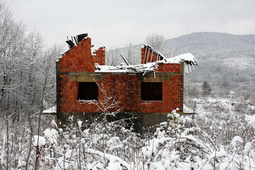 A partially collapsed red brick house stands deserted in a snow-covered field, surrounded by...