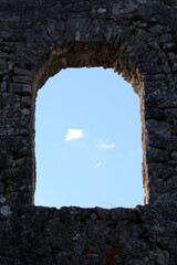 A rough-hewn stone window arch in ancient ruins frames a patch of blue sky, its weathered stones...
