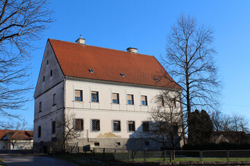 A large, rectangular manor house with a steep red tiled roof and barred lower windows stands in a...