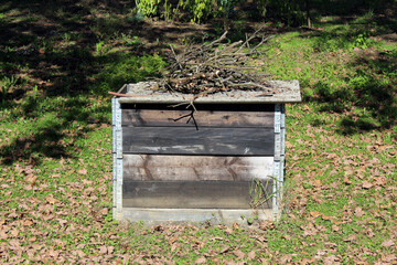 A square, homemade compost bin built from weathered wood and metal brackets sits on a leafy lawn,...