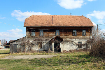 A large, two-story farmhouse with a tiled roof and wooden upper floor stands neglected, its facade...