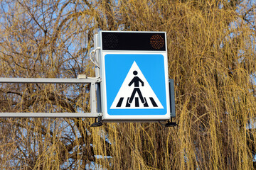 A blue and white pedestrian crossing sign with overhead amber warning lights is mounted on a metal...
