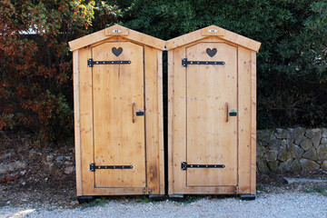Two side-by-side wooden outhouses with heart-shaped ventilation cutouts stand on gravel, backed by...