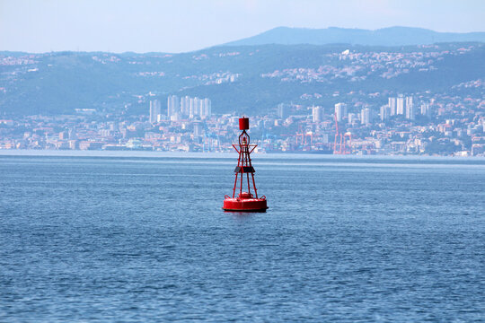 A bright red navigation buoy floats on tranquil blue water, with a distant city skyline, cranes, and forested hills visible across the bay under a clear sky