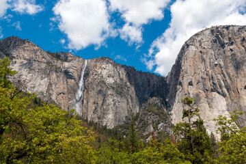 Yosemite Valley waterfall cascades down a cliff face under a blue sky with clouds.