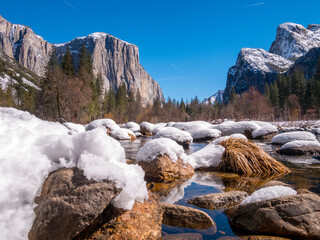 Snowy rocks in a stream with mountains and a clear blue sky in the background.